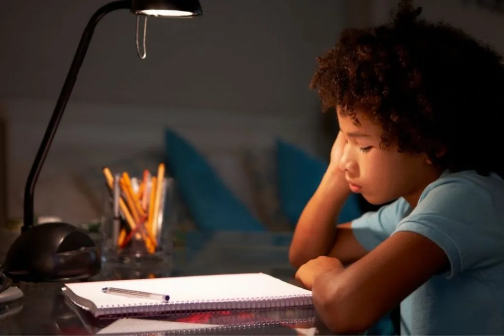 Child studying at night under a desk lamp, looking tired